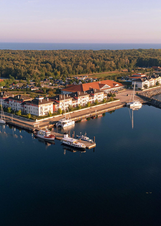 Luftaufnahme einer Marina mit Booten und einem angrenzenden Hotel, umgeben von Wasser und grüner Landschaft.