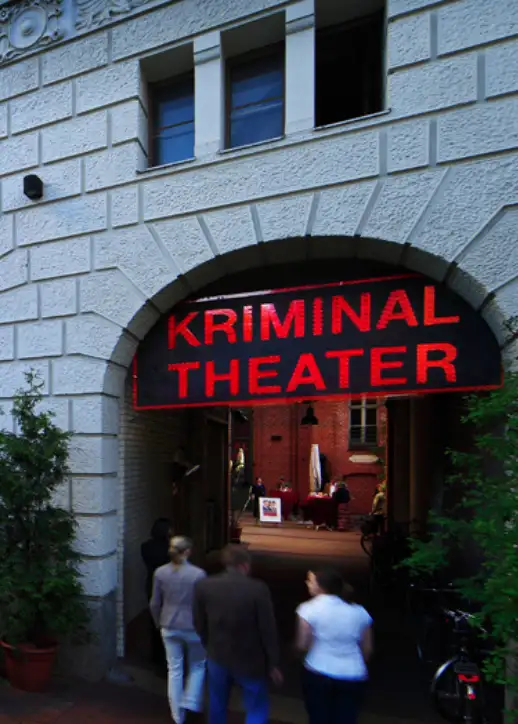 Ein Gebäude mit einem auffälligen Schild, das "Kriminaltheater" anzeigt, in einer belebten Stadtlandschaft.