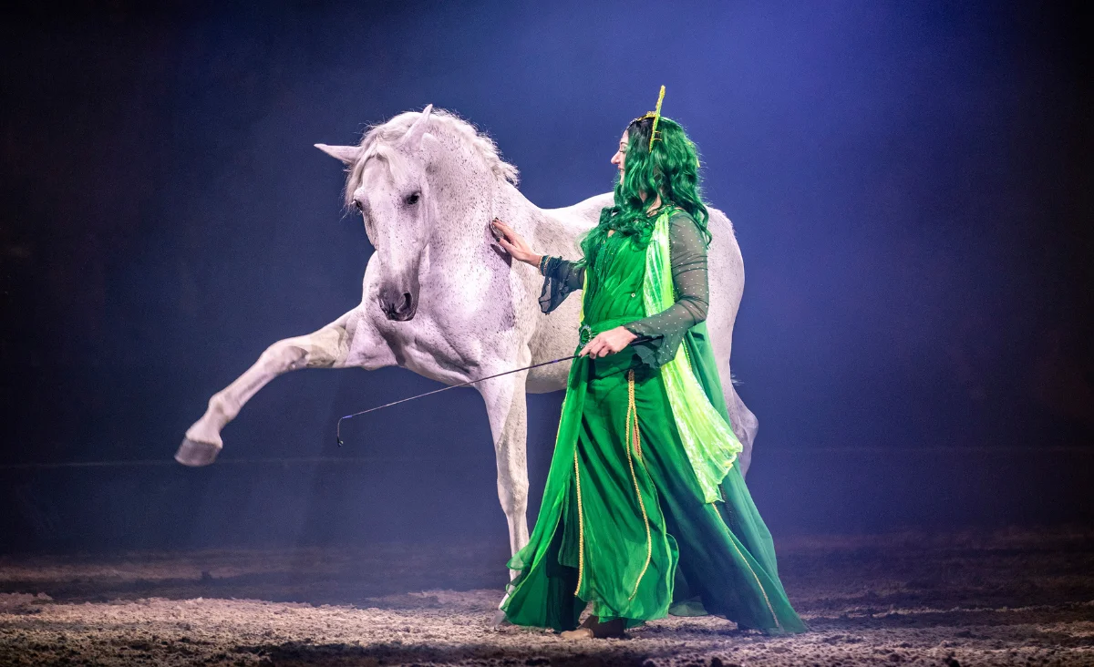 Eine Frau in grünem Kleid mit langen Haaren hält ein weißes Pferd in einer magischen Kulisse der CAVALLUNA-Show.