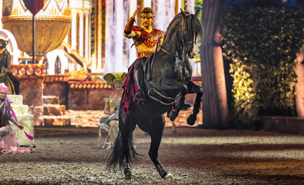 Eine Frau reitet auf einem Pferd in einer Zirkusaufführung, umgeben von bunten Lichtern und jubelndem Publikum.