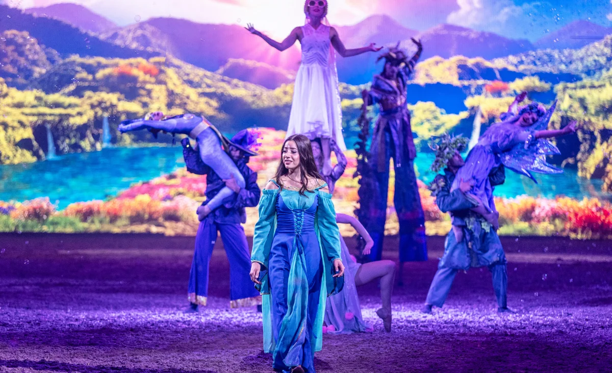 Eine Frau in einem blauen Kleid steht auf der Bühne, umgeben von anderen Darstellern während der CAVALLUNA-Show in Berlin.