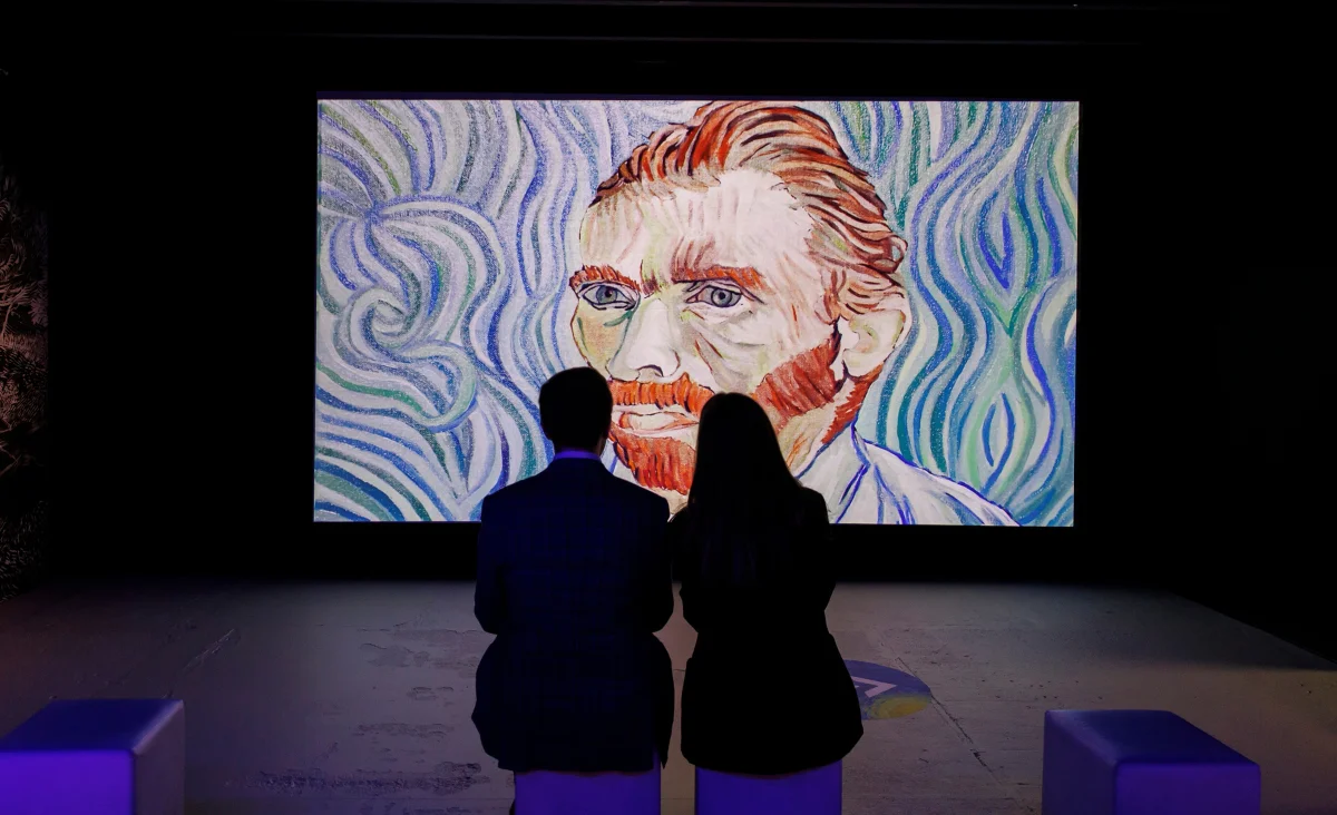 Zwei Personen sitzen vor einem großen Bildschirm mit einem Gemälde von Van Gogh in einer immersiven Ausstellung.