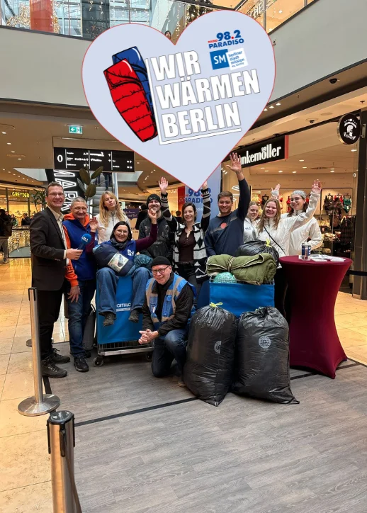 Eine Gruppe von Menschen posiert mit Gepäck in einem Einkaufszentrum zur Unterstützung der Aktion 'Wir wärmen Berlin 2025'.