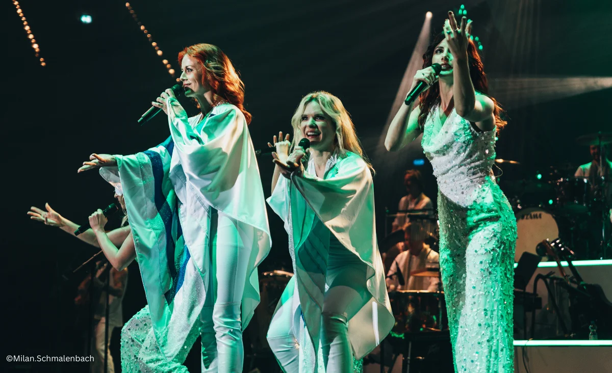 Drei Frauen in weißen Outfits stehen auf der Bühne mit Mikrofonen während ABBAMANIA THE SHOW in der Uber Arena Berlin.