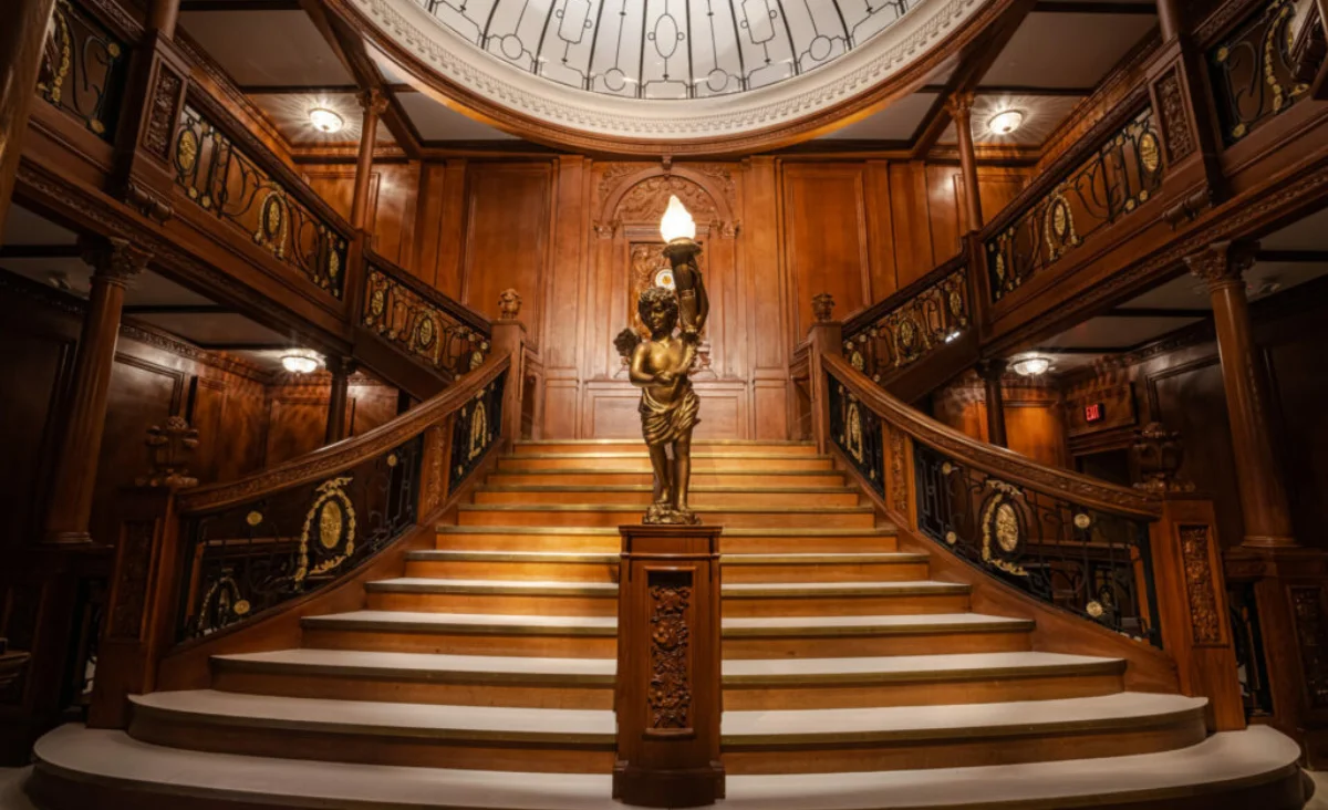 Große, reich verzierte Holztreppe mit goldener Statue einer Frau mit Fackel in der Mitte, umgeben von dunklem Holzvertäfelung und dekorativen Geländern in der Titanic-Ausstellung Potsdam