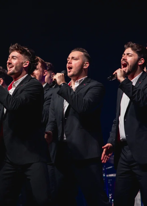 Gruppe von Sänger in schwarzen Anzügen mit weißen Hemden, die auf einer Bühne singen und Mikrofone halten