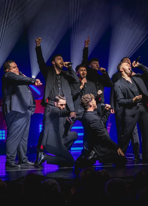 Mehrere Sänger in schwarzen Anzügen auf einer Bühne in dynamischen Posen mit erhobenen Armen und Mikrofonen: The 12 Tenors Konzert Berlin