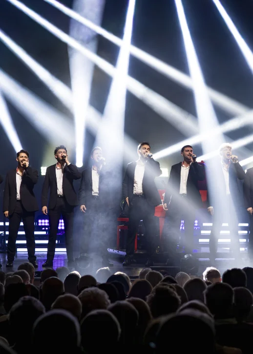 Sieben Männer in dunkler Kleidung stehen auf einer Bühne mit blauem Bühnenlicht und Scheinwerfern im Hintergrund