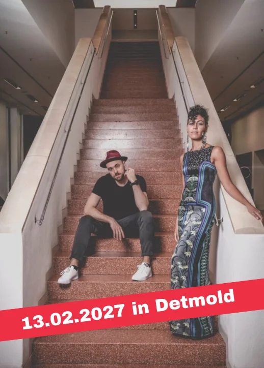 Ein Mann sitzt auf einer Treppe mit roten Stufen und trägt schwarze Kleidung und weiße Schuhe, eine Frau steht rechts neben der Treppe in einem langen, gemusterten Kleid