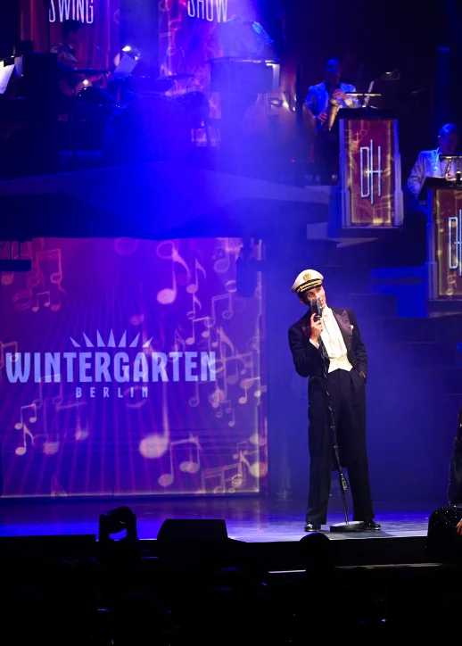 Sänger mit Matrosenmütze und Anzug steht vor Mikrofon auf Bühne mit beleuchtetem Wintergarten Berlin Schriftzug und Musiknoten im Hintergrund