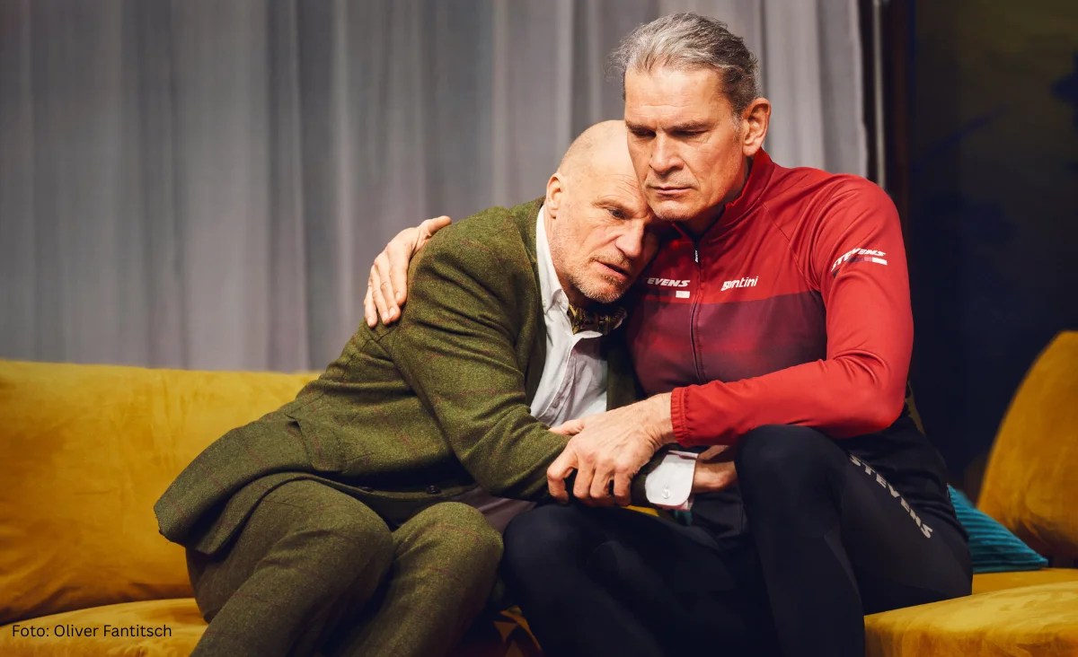 Zwei Personen sitzen eng nebeneinander auf einem gelben Sofa, eine Person legt den Arm tröstend um die andere und hält deren Hand