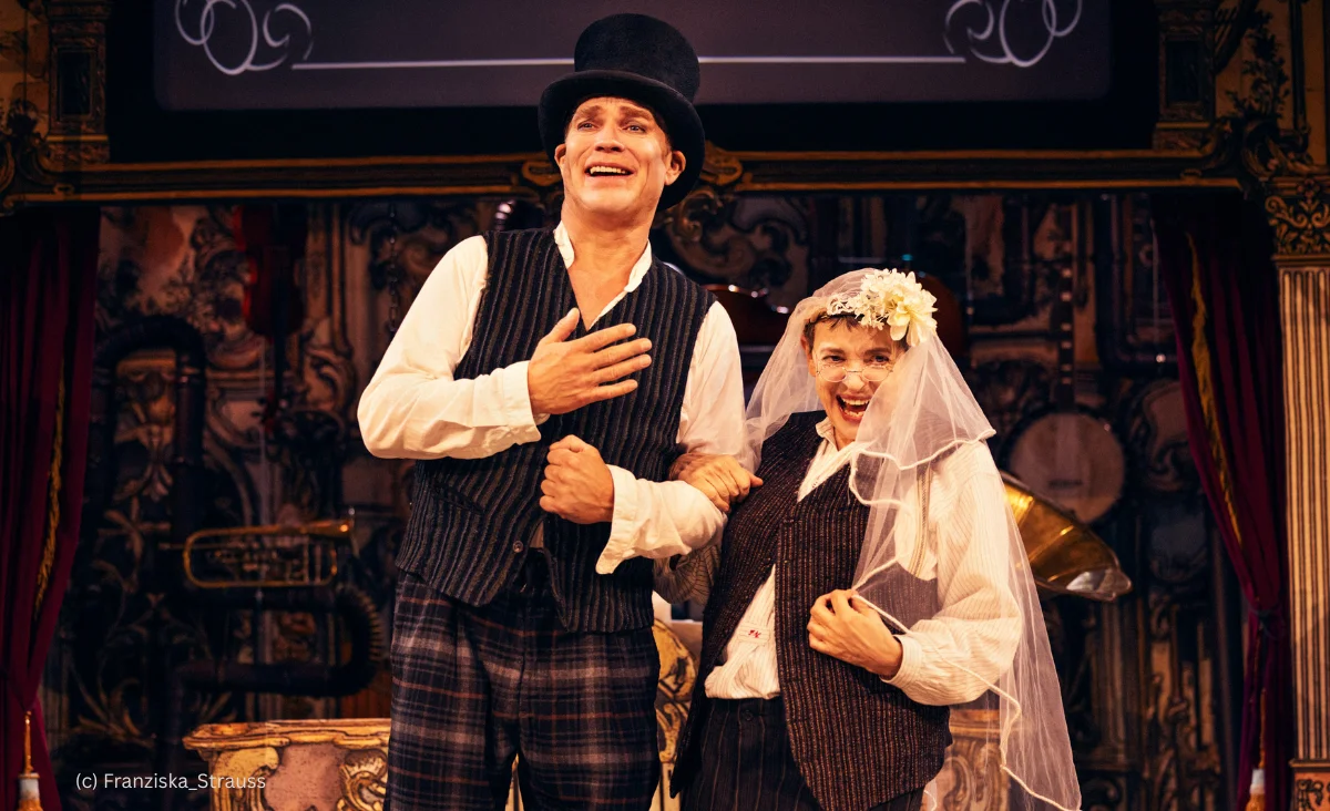 Zwei Personen in historischer Kleidung, eine mit Zylinder, die andere mit Schleier und Blumenkranz, stehen vor einer verzierten Wand und halten sich am Arm