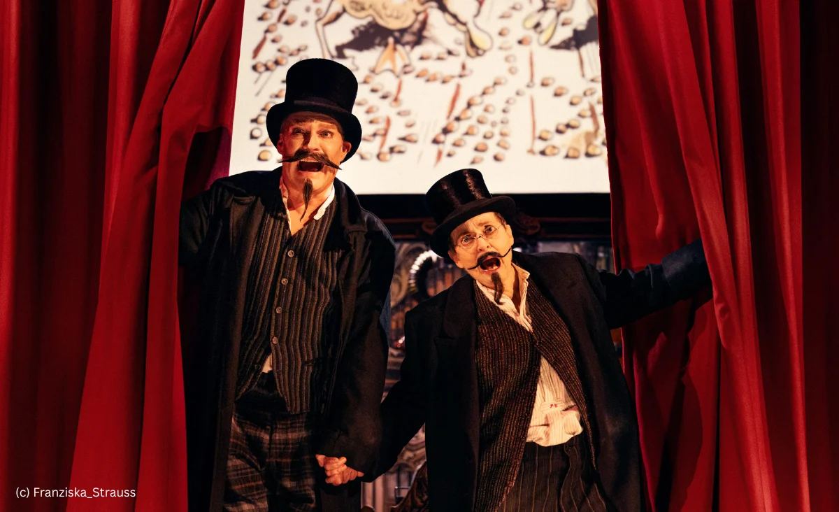 Zwei Männer in Anzügen und Zylindern stehen vor roten Vorhängen und halten sich an den Händen