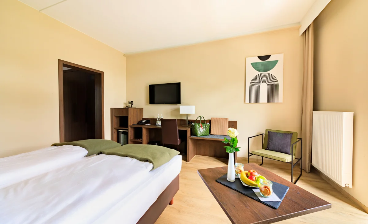 Hotelzimmer mit Doppelbett, Schreibtisch, Fernseher, Sessel, Tisch mit Obst und Vase sowie moderner Wanddekoration in einem Wellnesshotel in Bad Steben