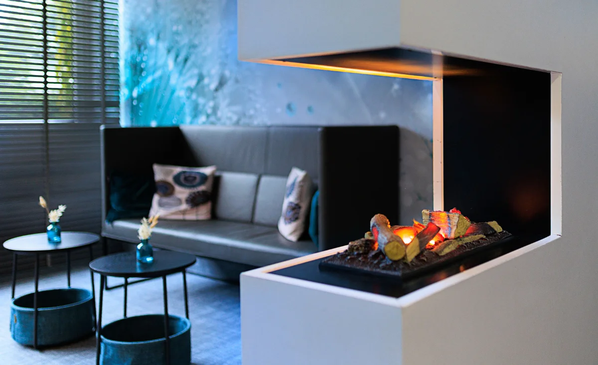 Moderner offener Kamin mit brennenden Holzscheiten vor grauem Sofa mit gemusterten Kissen und zwei kleinen runden Beistelltischen mit blauen Vasen