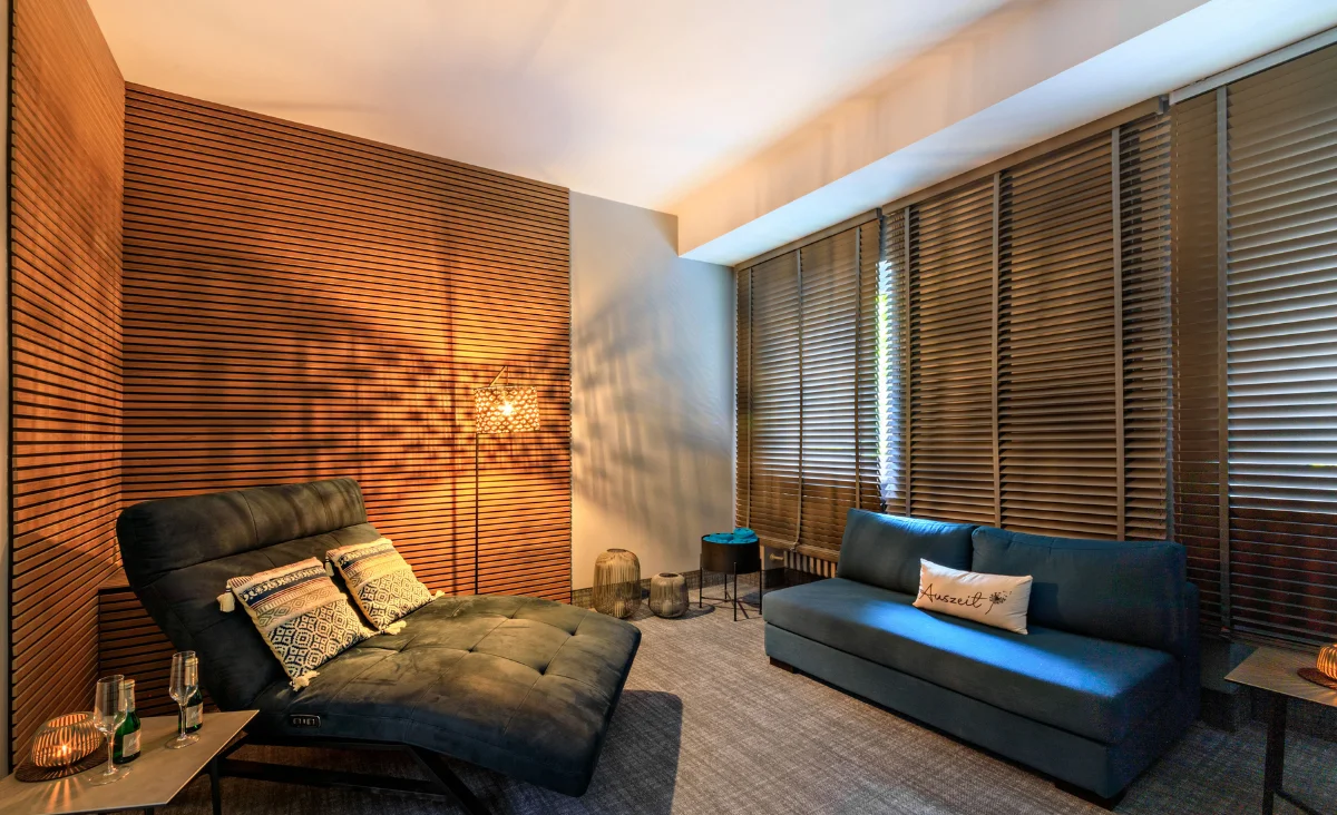 Gemütlicher Wellnessraum mit schwarzem Liegesessel, blauem Sofa, Holzwand mit Lichtmuster und großen Fenstern mit Jalousien im Wellnesshotel Bad Steben