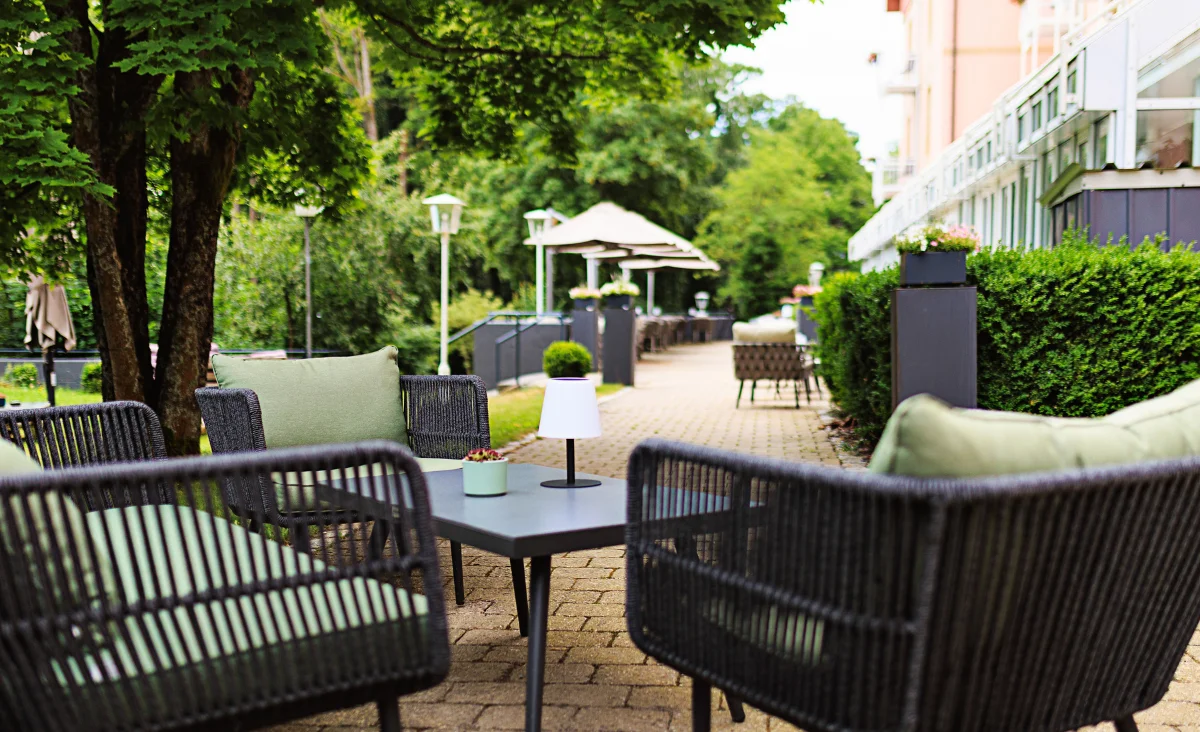 Terrasse mit Sitzgruppe aus Korbmöbeln, Tisch mit kleiner Lampe und Pflanzen, umgeben von Bäumen und Hecken neben einem Hotelgebäude