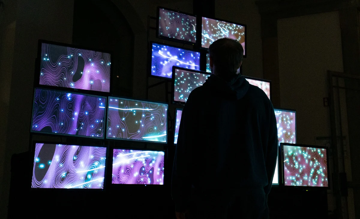 Person steht vor einer Wand aus mehreren Bildschirmen mit abstrakten, leuchtenden Mustern in der immersiven Ausstellung Nebula