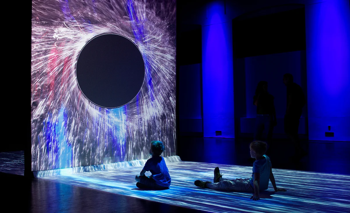 Zwei Kinder sitzen auf dem Boden vor einer großen, kreisförmigen Projektion mit strahlenförmigen Lichtmustern in einer dunklen Ausstellungsumgebung.