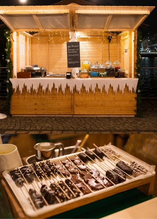 Collage vom All inclusive Weihnachtsmarkt an der Spree in Berlin: Ein festlich beleuchteter Holzstand mit Süßwaren am Spreespeicher im oberen Bereich, kombiniert mit einer Nahaufnahme von frischen Schokofrüchten am Spieß im unteren Bereich.