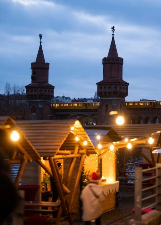 Blick auf den All inclusive Weihnachtsmarkt an der Spree in Berlin mit den beleuchteten Holzhütten am Spreespeicher und der Oberbaumbrücke im Hintergrund, überquert von einer gelben U-Bahn.