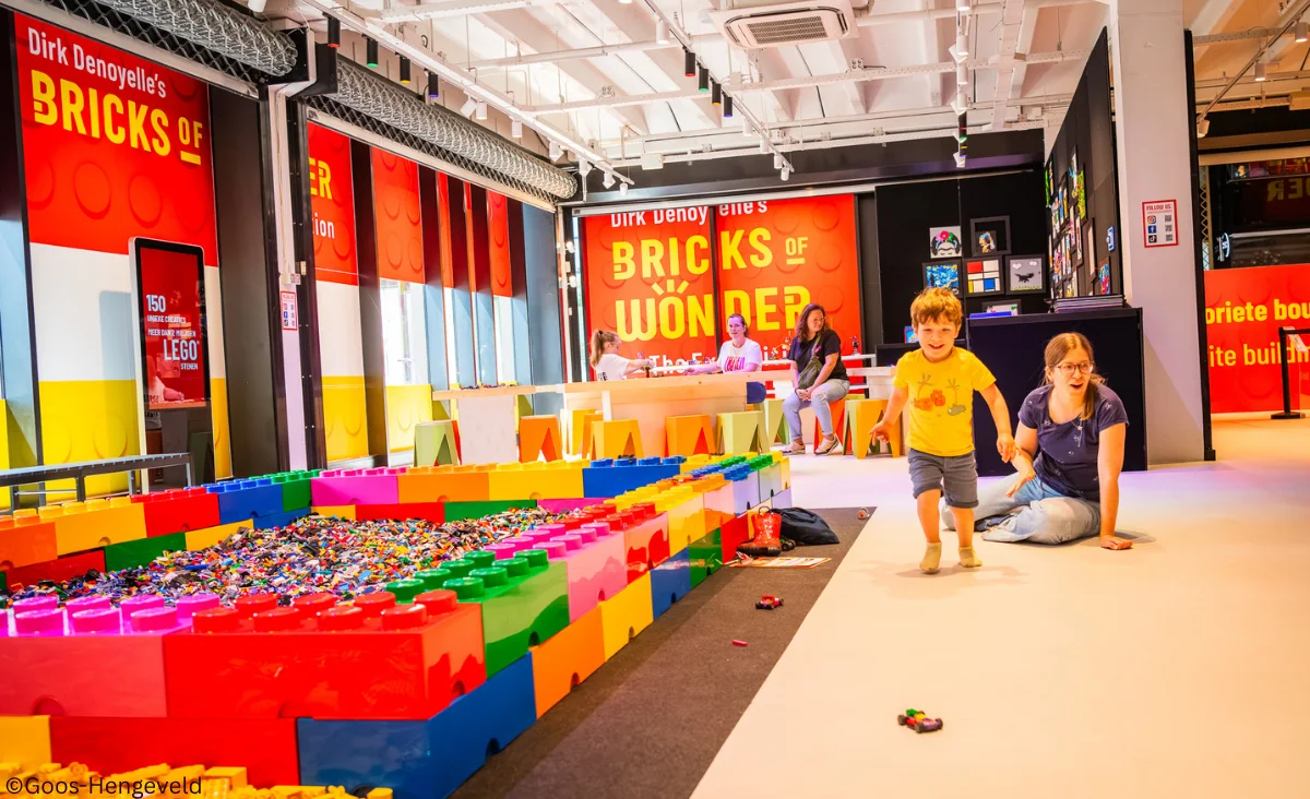 Ein Kind läuft auf einem hellen Boden vor einer Frau sitzend neben einem großen, bunten Lego-Bau mit großen Steinen in einem Ausstellungsraum von Bricks of Wonders.