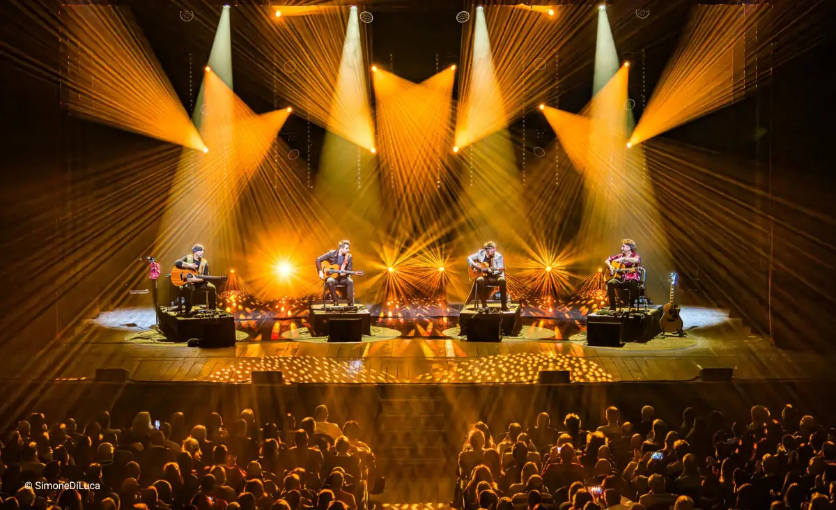 Vier Musiker sitzen auf Stühlen auf einer Bühne mit akustischen Gitarren und werden von mehreren gelben Scheinwerfern beleuchtet während der Gitarrensensation 40 Fingers.