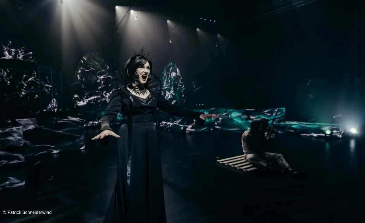 Schauspielerin in schwarzem Kostüm mit dramatischem Gesichtsausdruck auf dunkler Bühne mit abstrakten Lichtprojektionen und einer Person, die auf einem Holzgestell liegt während der "This is The Greatest Show Tour".