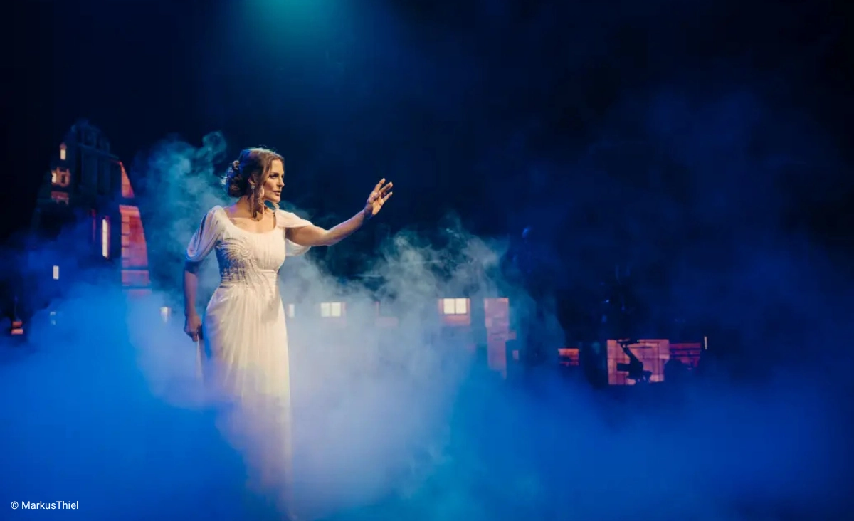Frau in weißem Kleid steht auf nebliger Bühne mit dunklem Hintergrund und hebt eine Hand während der "This is The Greatest Show Tour".