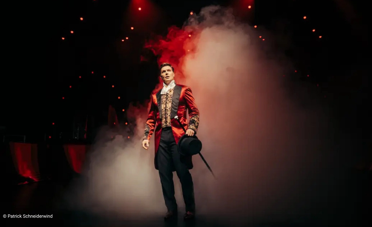 Ein Mann in rotem Frack mit schwarzem Zylinder und Stock steht vor einer Nebelwolke auf einer Bühne mit roten Lichtern im Hintergrund während der "This is The Greatest Show Tour".