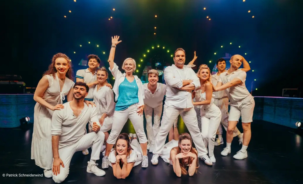 Gruppe von elf Personen in weißer Kleidung posiert auf einer Bühne mit dunklem Hintergrund und bunten Lichtringen während der "This is The Greatest Show Tour".
