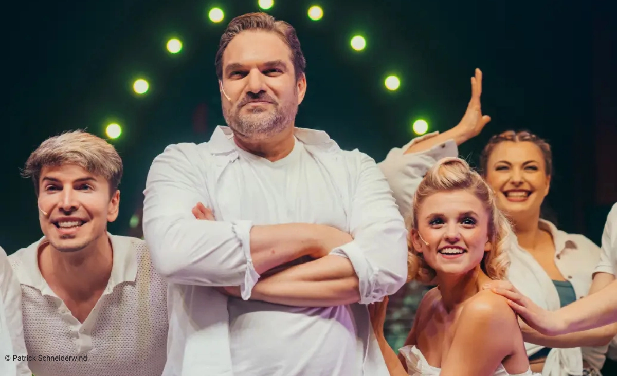 Vier Schauspieler in Bühnenkostümen posieren vor einem dunklen Hintergrund mit grünen Lichtern, der mittlere Mann steht mit verschränkten Armen, die anderen lächeln und posieren lebhaft während der "This is The Greatest Show Tour".