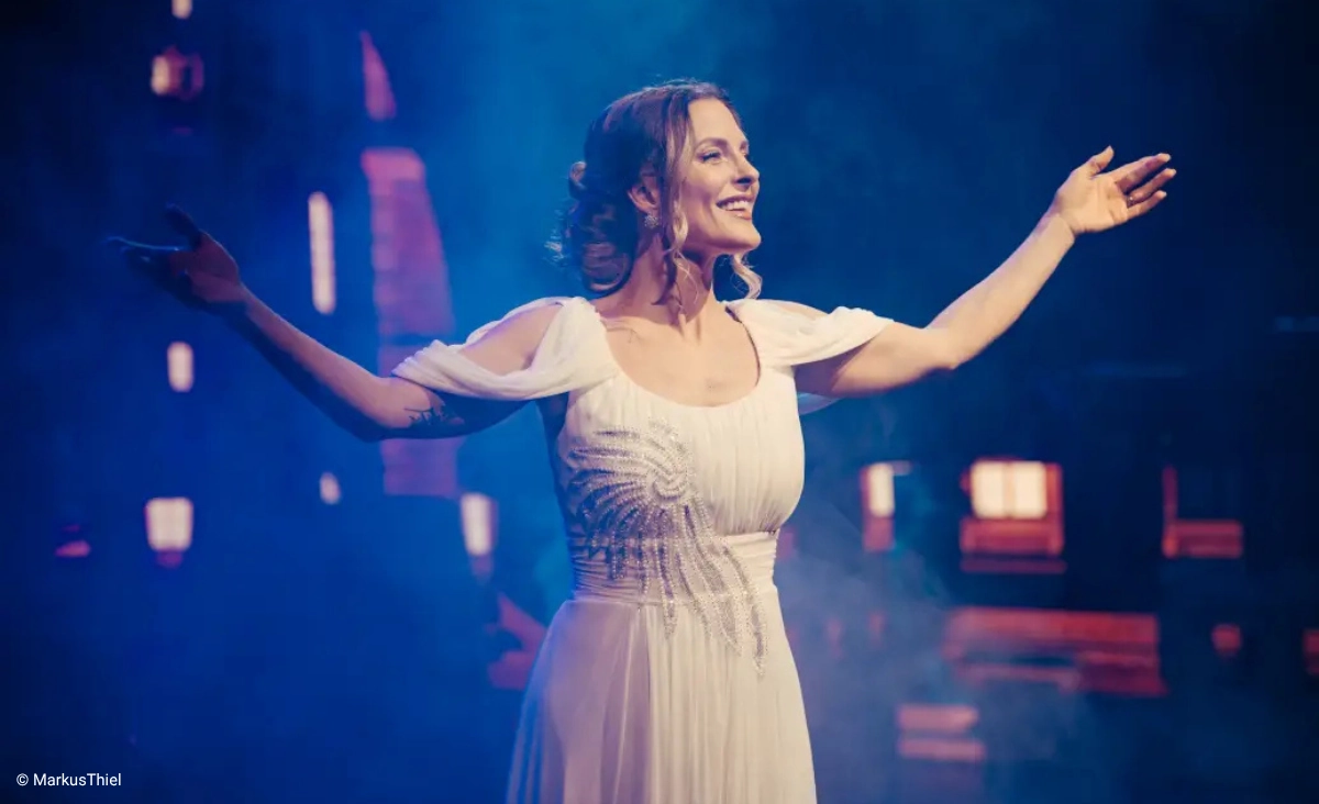 Frau in weißem Kleid steht auf nebliger Bühne mit dunklem Hintergrund und hebt eine Hand während der "This is The Greatest Show Tour".