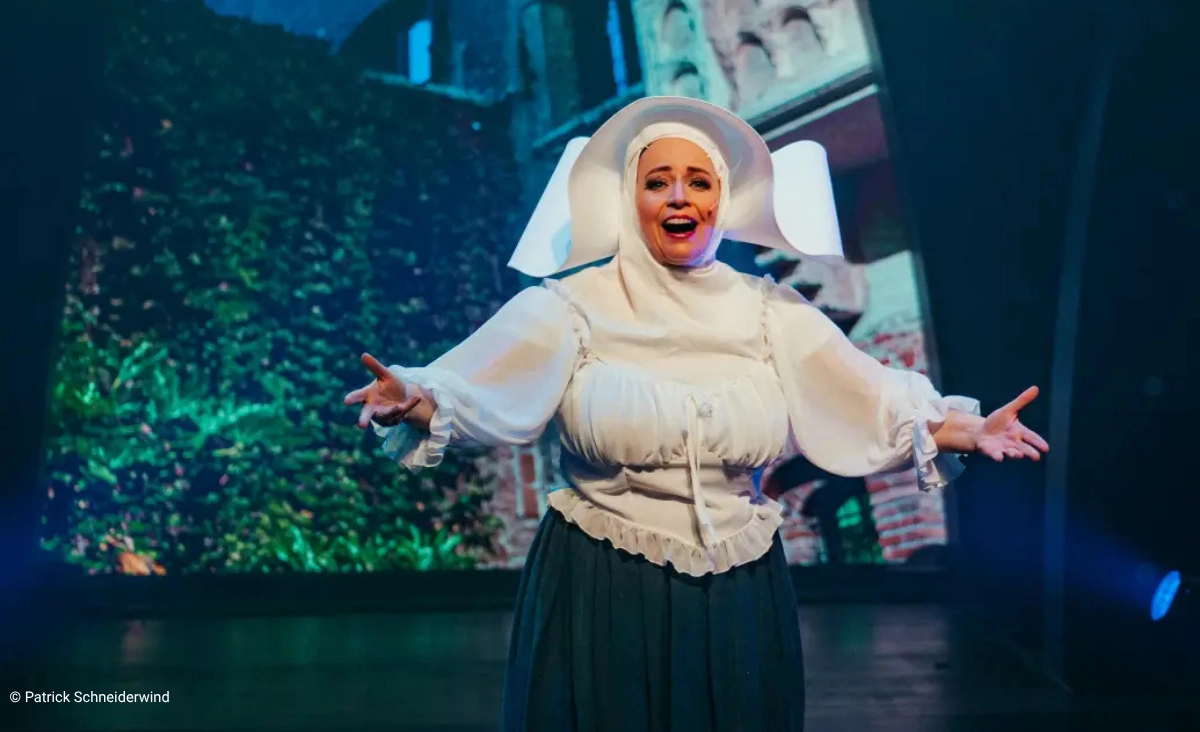 Schauspielerin in historischer Nonnenkleidung mit ausgebreiteten Armen auf einer Bühne vor einer Kulisse mit Gebäudefassade und Efeu während der "This is The Greatest Show Tour".