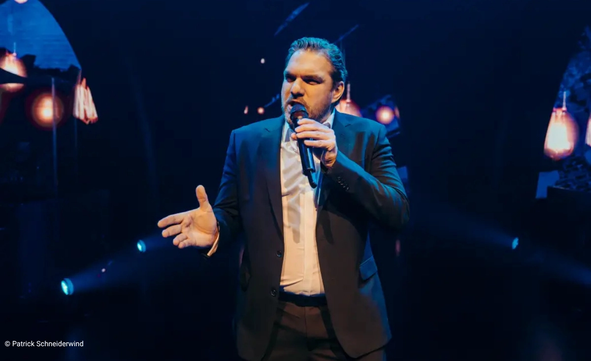 Mann in dunklem Anzug mit Mikrofon auf Bühne vor dunklem Hintergrund mit blauen und orangen Lichtern während der "This is The Greatest Show Tour".