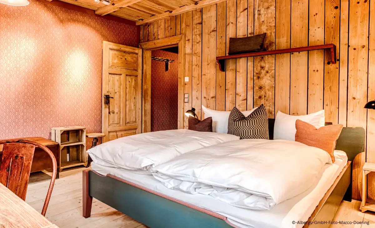 Gemütliches Hotelzimmer mit Holzwänden, Doppelbett mit weißen Decken und Kissen in verschiedenen Farben, Holztür und Stuhl in rustikalem Stil