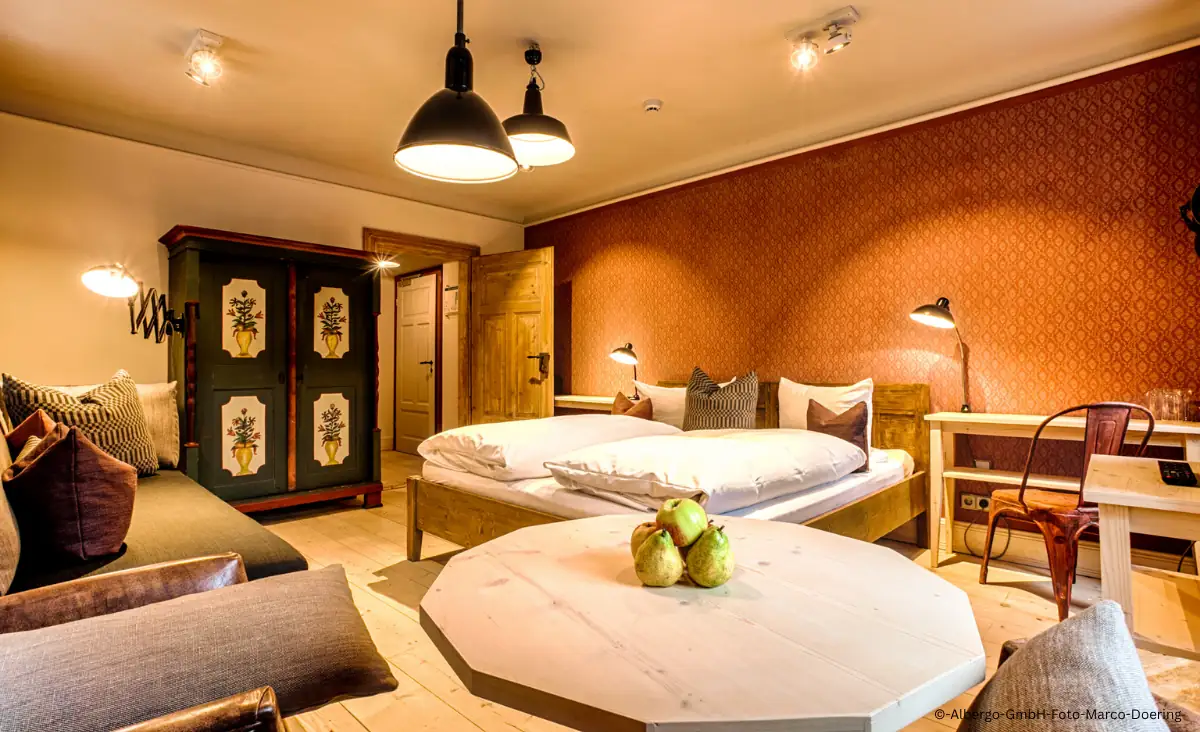 Gemütliches Hotelzimmer mit Doppelbett, Holzmöbeln, einem bemalten Schrank, einem runden Tisch mit drei grünen Äpfeln und warmem Licht im Winterdorf Schmilka Hotel Zur Mühle